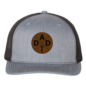 May include: Grey trucker cap with a brown circular patch displaying the word "DAD". The cap has a black mesh back and a curved peak. The patch also features a graphic of a utility pole.
