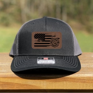 May include: A black and gray trucker hat with a curved brim. The hat features a brown leather patch with a distressed American flag and a fire department emblem. The hat has a Richardson 112 tag.