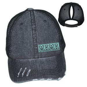 May include: A distressed black trucker hat with a mesh back and a curved brim. The front of the hat features the word "MAMA" in a teal rectangular design. The hat has a ponytail opening.
