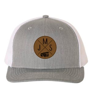 May include: Grey and white trucker cap with a brown leather patch. The patch features the letters "JMS" crossed with a fire engine graphic. The cap has a curved peak and a mesh back.