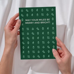 May include: A greeting card held by hands, with a dark green background and white icons of running shoes, watches, runners, water bottles, and trees. The card reads "MAY YOUR MILES BE MERRY AND BRIGHT!"