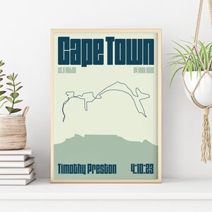 May include: A framed print featuring the words "Cape Town" in dark blue. Below, a line drawing of a mountain range and the text "Timothy Preston 4:10:23". The background is light green and blue.