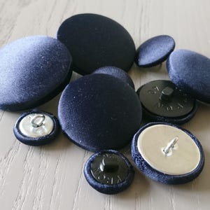 May include: A collection of nine navy blue buttons in different sizes and styles. Some buttons have a silver shank, while others have a black plastic shank.