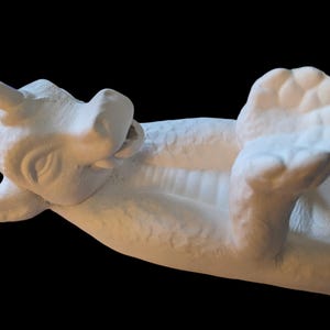 May include: White ceramic incense burner shaped like a dragon lying on its side with its head turned to the side and its mouth open. The dragon has a horn on its head and its wings are spread out.