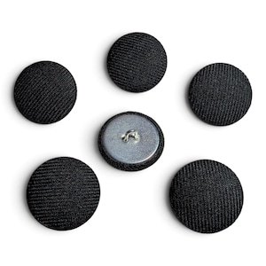 May include: A set of five circular buttons covered in a black fabric with a visible woven texture. Four buttons are positioned with the fabric side facing up, while one central button is flipped to show the silver metal back with a wire shank.