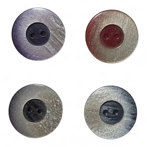 Può includere: Quattro bottoni rotondi con finitura metallica. I bottoni hanno un centro scuro con due fori. L'anello esterno dei bottoni ha un aspetto strutturato e iridescente in tonalità argento, grigio e bronzo.