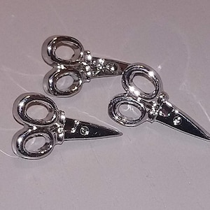 Könnte beinhalten: Zwei silberfarbene Metall-Schlüsselanhänger in Form von Scheren. Die Anhänger sind detailliert und haben ein realistisches Aussehen.