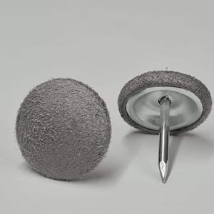 Op de afbeelding: Twee grijze stoffen meubelstiften met een zilveren spijker. Eén stift is van bovenaf te zien en de andere van de zijkant.