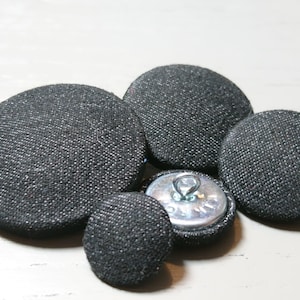 May include: Five black fabric covered buttons with a silver metal shank. The buttons are round and have a textured surface.