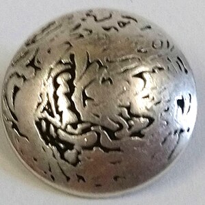 May include: A silver-toned button with a textured, abstract design. The button has a raised, rounded shape.