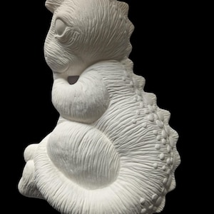 May include: A white ceramic bisque figurine of a seated dinosaur with a textured surface. The creature features a prominent dorsal fin with rounded spikes running down its back, a rounded belly, and a smooth, unpainted matte finish throughout.