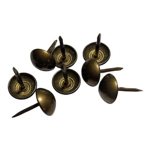 May include: A selection of antique brass upholstery tacks. Each tack has a rounded, domed head and a long, slender nail. The heads have a warm, metallic finish, and the nails are pointed for easy insertion into fabric or wood.