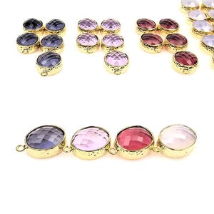May include: An assortment of gold-toned bezel-set gemstone pendants in various colors, including purple, pink, and red. The gemstones have a faceted cut, and the pendants are arranged on a white background.