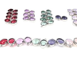 May include: An assortment of oval-shaped gemstone pendants in various colors, including red, pink, purple, green, and blue. Each pendant has a silver-colored bezel and a small loop for attaching to a chain or other jewelry component. The gemstones are faceted and have a polished finish.