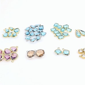 May include: An assortment of gemstone jewelry components in various shapes and colors. The components include light blue, pink, and clear stones set in gold and silver-toned metal settings. The components are arranged on a white surface.
