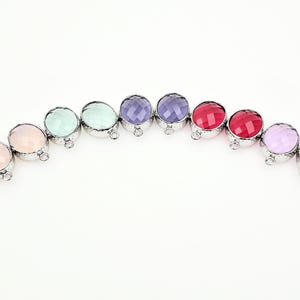 May include: A row of ten circular charms, each featuring a faceted, translucent gemstone set in a silver-toned metal frame with a small loop at the bottom. The stones display a variety of colors including pale pink, mint green, purple, and red.