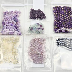 May include: Assortment of gemstone beads in clear plastic bags. The beads are various shades of purple, lavender, and gold. The beads are in different shapes and sizes, including round and faceted.