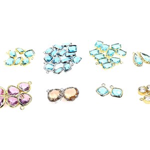 May include: A collection of faceted glass charms in various shapes and colors, including light blue, pink, and champagne, set in gold-toned metal frames. Each charm features small loops for attachment, arranged in groups across the frame.