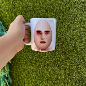 Könnte beinhalten: Ein weißer Keramikbecher mit einem grafischen Aufdruck eines menschlichen Gesichts, das auf ein Ei projiziert ist. Das Gesicht hat dunkles Haar, dunkle Augenbrauen und rötlich getönte Augen mit einem neutralen Ausdruck. Der Becher hat einen Henkel.