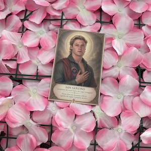 Peut inclure: Une carte de prière rectangulaire illustrant un homme avec une auréole, portant une chemise vert foncé avec un écusson circulaire, posée sur un fond de fleurs artificielles roses et blanches. Le texte indique SAN SANTINO.