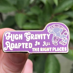 May include: Purple enamel pin with a gold outline. The pin features a cartoon creature with the text "High Gravity Adapted In All The Right Places".