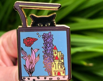 May include: A gold-colored enamel pin depicting a black cat peeking into a fish tank. The tank contains a red fish, purple plants, and a yellow castle.