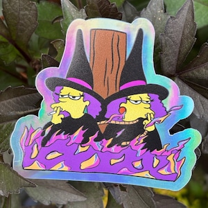May include: Holographic sticker featuring two cartoon witches with yellow skin, purple hair, and black hats with pink bands. They are smoking and surrounded by purple flames. The background is a blurred image of green leaves.