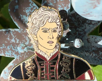 May include: Gold-toned enamel pin featuring a portrait of a person with white hair, wearing a black and red patterned jacket with gold accents. The pin is set against a blue and gray background.