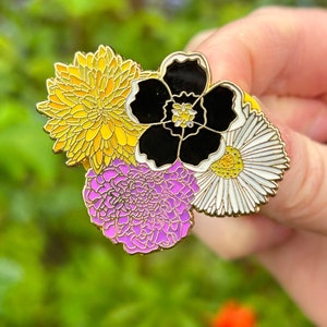 Op de afbeelding: Een gouden emaille pin met vier bloemen in een cluster. De bloemen zijn geel, zwart, wit en paars. De pin is een representatie van de non-binaire pride-vlag.