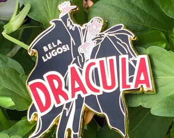 May include: Gold-colored enamel pin featuring a black and white illustration of Bela Lugosi as Dracula. The pin has a red and white "Dracula" text.