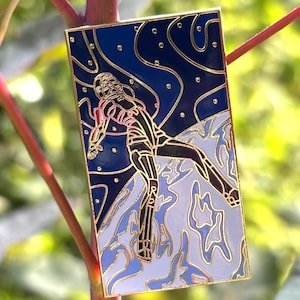 May include: Rectangular enamel pin featuring a stylized figure in a dynamic pose against a backdrop of blue and white abstract shapes. The pin has a gold border and accents, with the figure outlined in black and red.