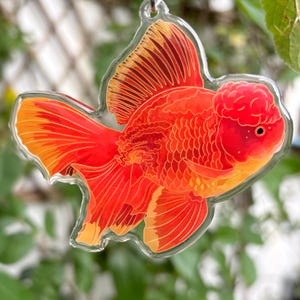 May include: A clear acrylic keychain featuring a vibrant orange and red goldfish design. The fish has detailed fins and a distinctive head shape, with a silver-colored metal ring for attachment. The background is blurred, suggesting an outdoor setting.
