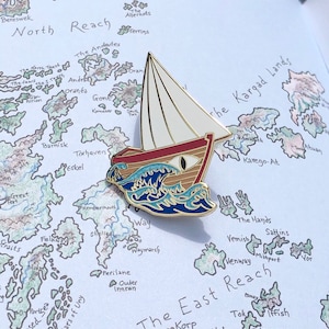 May include: A gold-coloured enamel pin depicting a boat with a white sail and a red hull, sailing on blue waves. The pin is set against a map background with handwritten text.