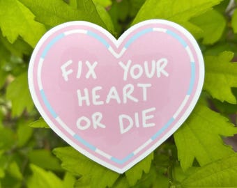May include: A pink heart-shaped sticker with the text "FIX YOUR HEART OR DIE" in white. The sticker has a light blue and white border and is set against a background of green leaves.