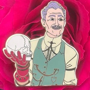 May include: A gold-tone enamel pin featuring a cartoon illustration of a man with purple hair, a white mustache, and a green vest holding a white skull. The man is smiling.