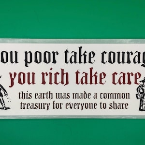May include: A rectangular sign with a white background and black and maroon text. The text reads "you poor take courage, you rich take care, this earth was made a common treasury for everyone to share." Two cartoon figures are on either side of the text.