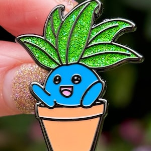 May include: A blue and green enamel pin of a cartoon plant with a happy face in a terracotta pot. The plant has green glitter leaves.
