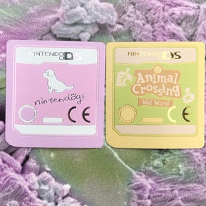 May include: Two Nintendo DS game cartridges. The left cartridge is pink and features a white dog with the text "nintendogs". The right cartridge is yellow and features a green leaf with the text "Animal Crossing Wild World".
