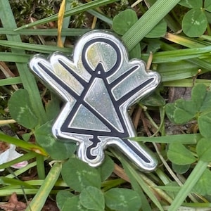 Puede incluir: Un pin de metal plateado con un contorno negro de una figura abstracta, sobre un fondo de hierba verde y tréboles. El pin presenta un diseño estilizado con brazos extendidos y una cabeza circular.