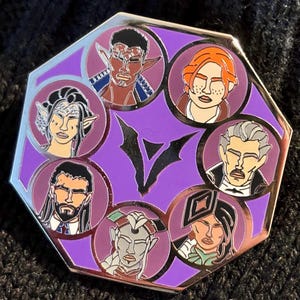 May include: A purple octagonal enamel pin with a stylised V in the centre. The pin features eight circular portraits of different characters with various skin tones, hair colours, and facial features.