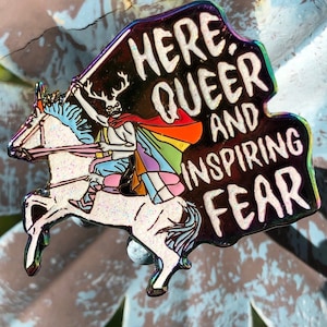 May include: A rainbow enamel pin with a black background and the text "Here. Queer and Inspiring Fear". A cartoon figure with antlers and a rainbow cape rides a white unicorn.