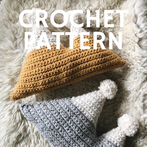 Op de afbeelding: Twee gehaakte kussens, één mosterdgeel en één grijs en wit, liggen op een zacht wit oppervlak. Het gele kussen is rechthoekig. Het grijs-witte kussen heeft de vorm van een narrenmuts met witte pompons. De tekst "CROCHET PATTERN" is overgelegd.