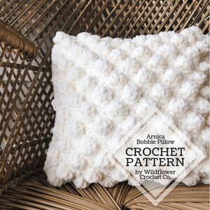 May include: White crochet pillow with a textured bobble stitch pattern. The pillow is sitting on a wicker chair. The text "Arnica Bobble Pillow CROCHET PATTERN by Wildflower Crochet Co." is on the pillow.