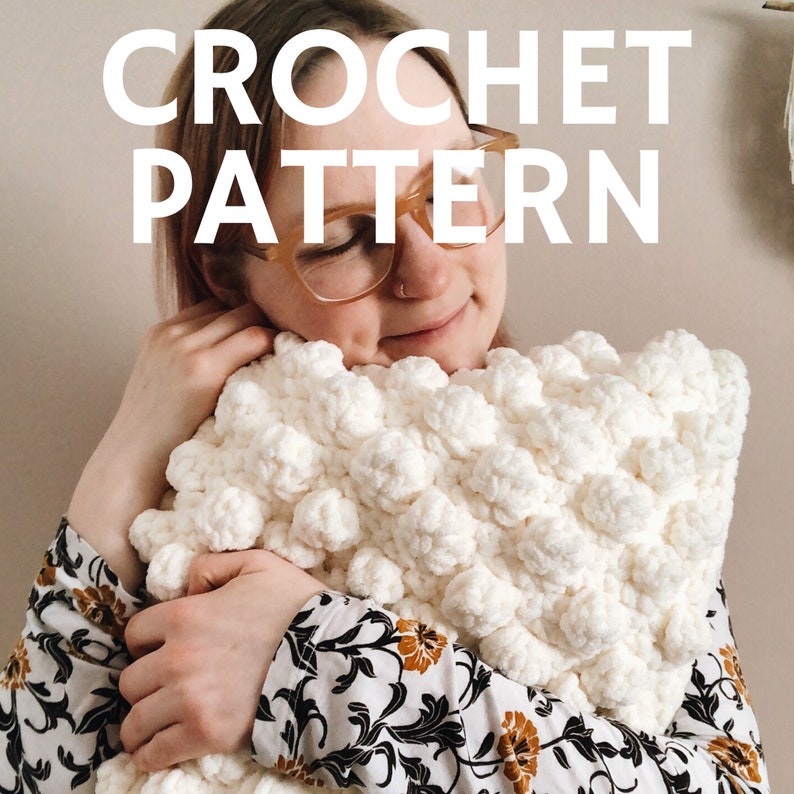 May include: A white crocheted pillow with a textured, puffy design. The pillow is being held by a person wearing a floral shirt. The text "CROCHET PATTERN" is displayed above the pillow.