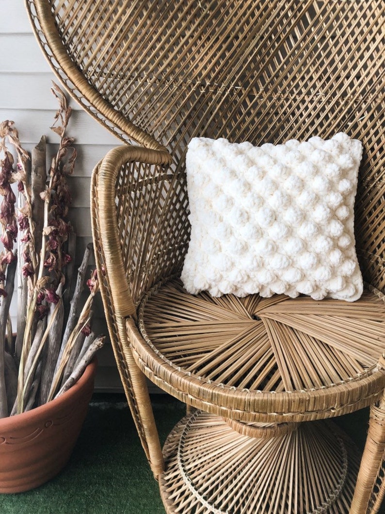 May include: A white crocheted pillow with a textured, puffy design sits on a wicker peacock chair. The chair has a round seat and a tall, curved back.