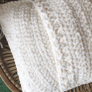 May include: A white crocheted pillow with a textured, fluffy surface. The pillow is folded in half and placed in a woven basket.