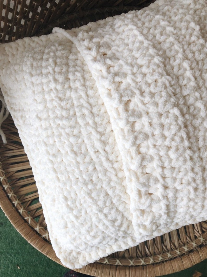 May include: A white crocheted pillow with a textured, fluffy surface. The pillow is folded in half and placed in a woven basket.