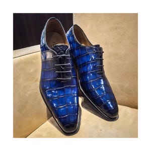 Peut inclure: Une paire de chaussures de ville en cuir bleu avec une texture peau de crocodile. Les chaussures ont un design à lacets et une finition brillante. Les chaussures sont présentées sur une surface de ton neutre.
