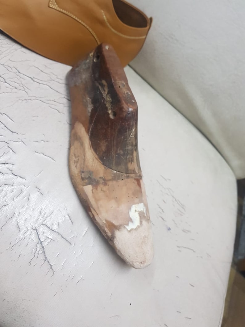 Puede incluir: Horma de zapato de madera usada, con una puntera de color claro. La horma es de color marr&oacute;n oscuro y tiene agujeros perforados en la parte superior. Un zapato de cuero marr&oacute;n claro es parcialmente visible en el fondo.