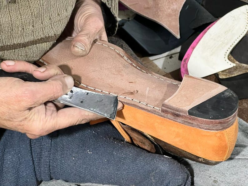 May include: A shoemaker uses a sharp knife to trim the edge of a leather shoe component. The shoe is tan and orange, with a black accent. The shoemaker is wearing a brown sweater and blue pants. Other shoe parts are in the background.